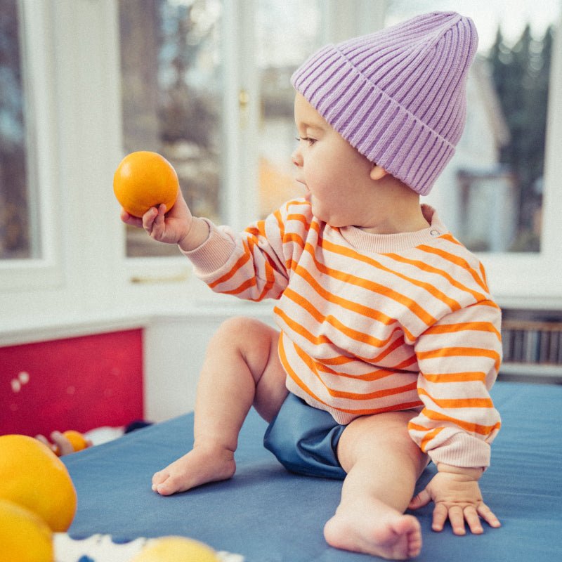 Baby in Judes Windel mit lila Mütze hält Mandarine und sitzt auf Tisch